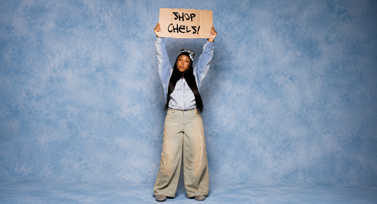 A woman stands against a mottled blue studio backdrop holding a cardboard sign above her head that reads 'SHOP CHELS!'. She is wearing a light blue denim shirt, a black top underneath, and wide-leg beige pants.